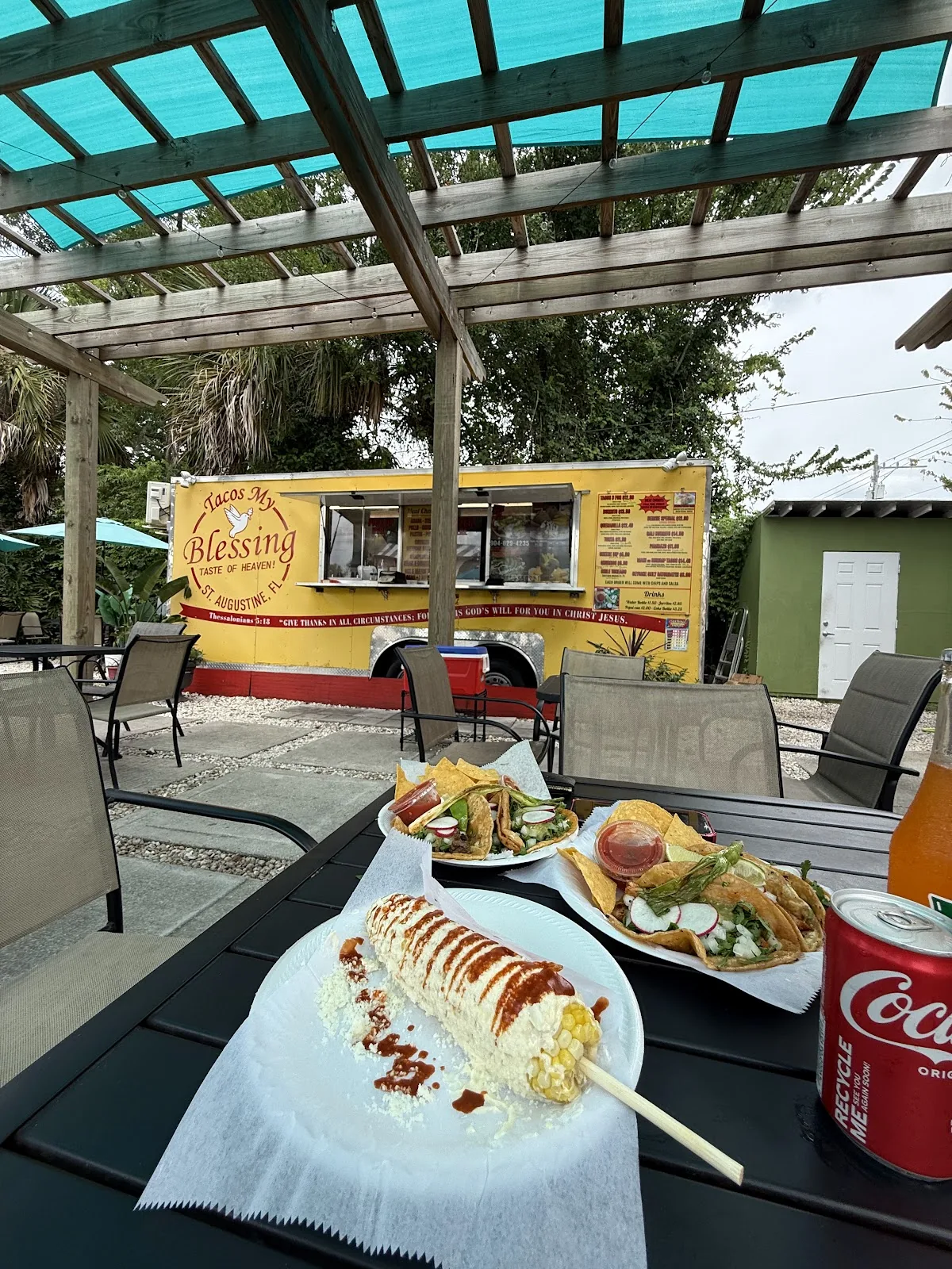 Tacos and elote on a patio table, with the yellow Tacos My Blessing food truck behind under a pergola.